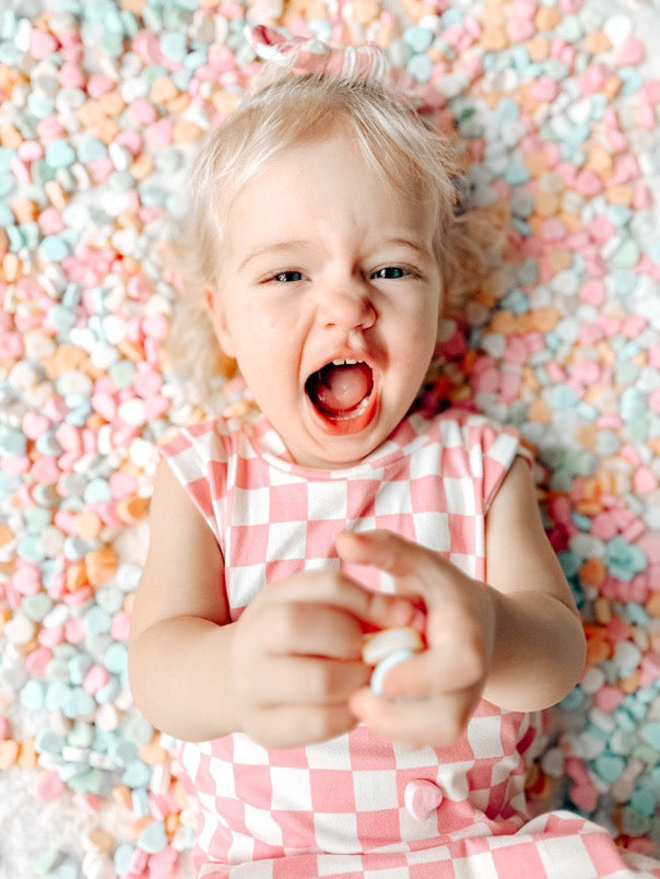 Toddler girl in pink checkered romper lying on pastel candy hearts – Adorable Valentine’s Day outfit for babies and toddlers. Soft and stylish checkered dress, perfect for festive photoshoots, playdates, and holiday celebrations. Cute and comfy handmade kids&