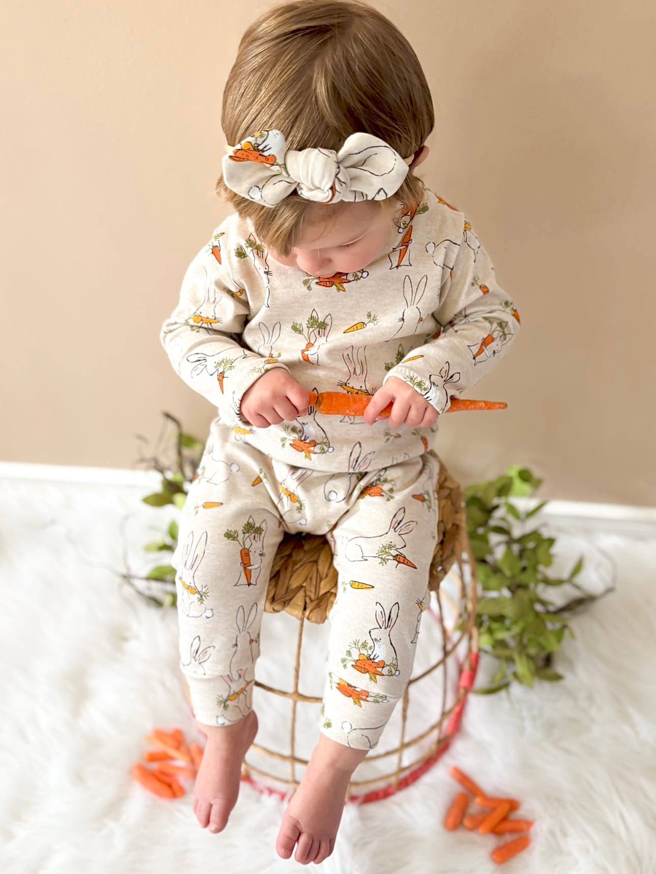 Adorable baby wearing an Easter bunny and carrot romper with a matching headband, sitting on a woven stool, perfect for a baby’s first Easter outfit.