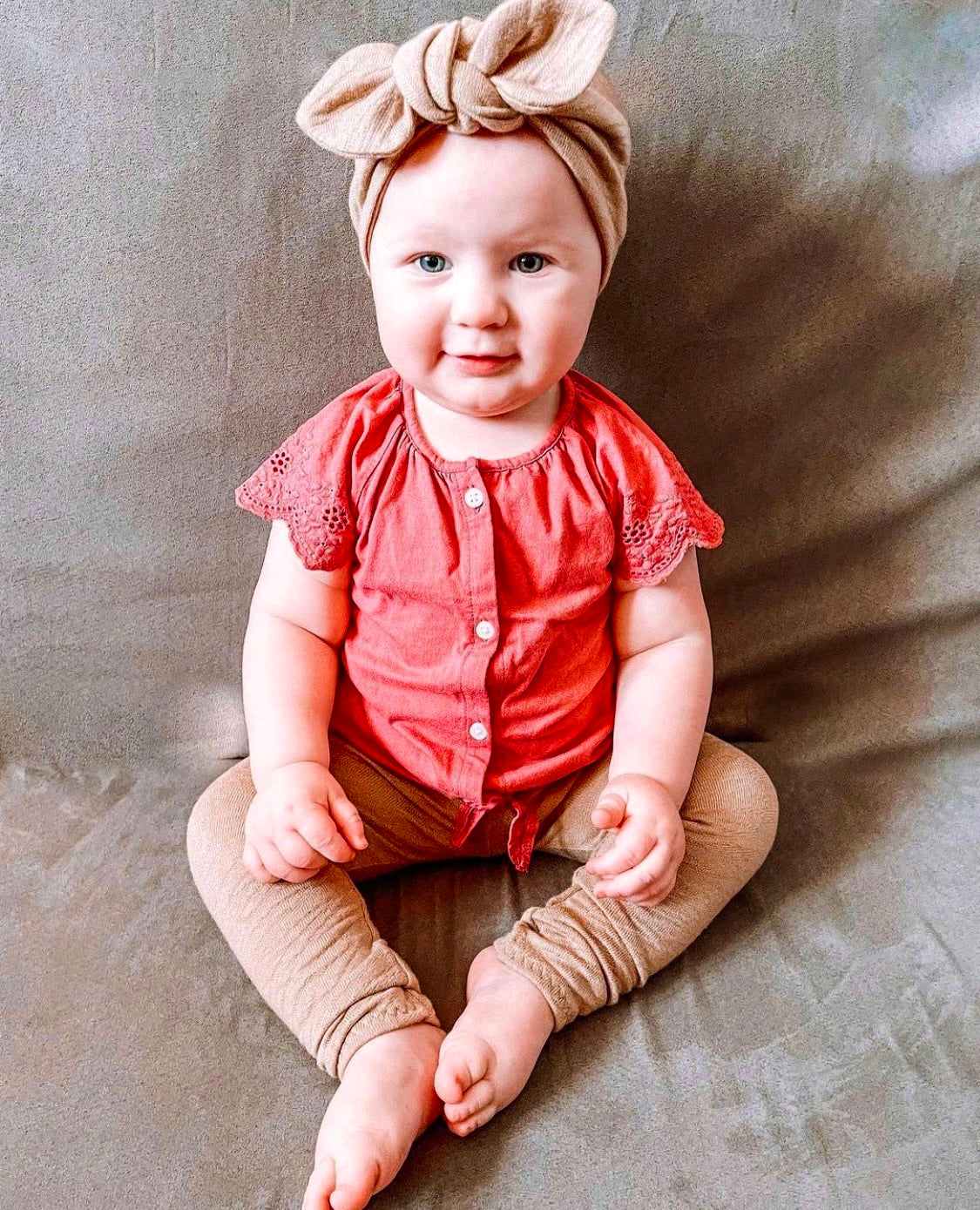 Baby wearing a red top and camel beige pants with a headband, sitting on a neutral background