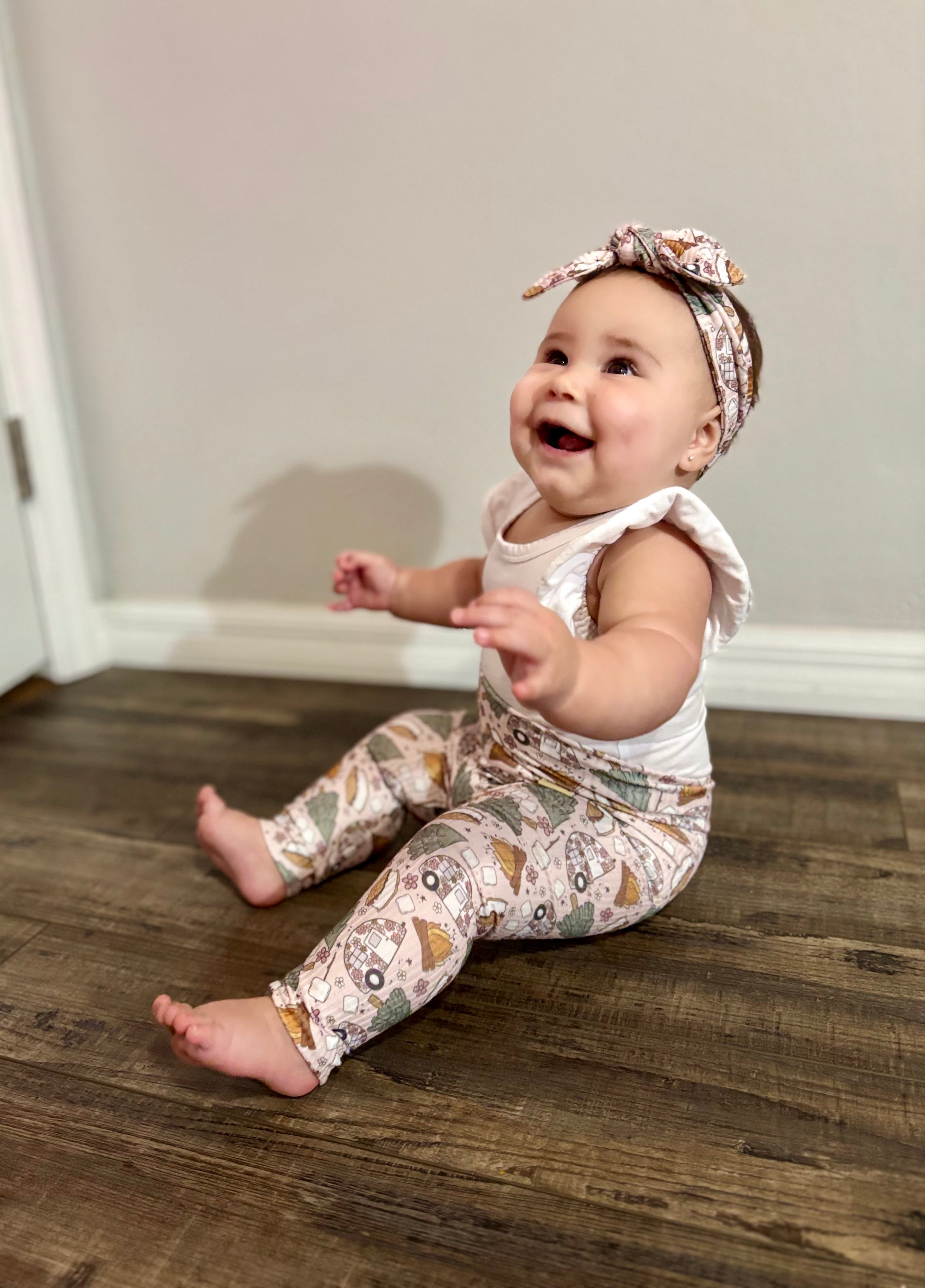 Baby sitting on a wooden floor wearing a white top and matching camping print leggings and headband.