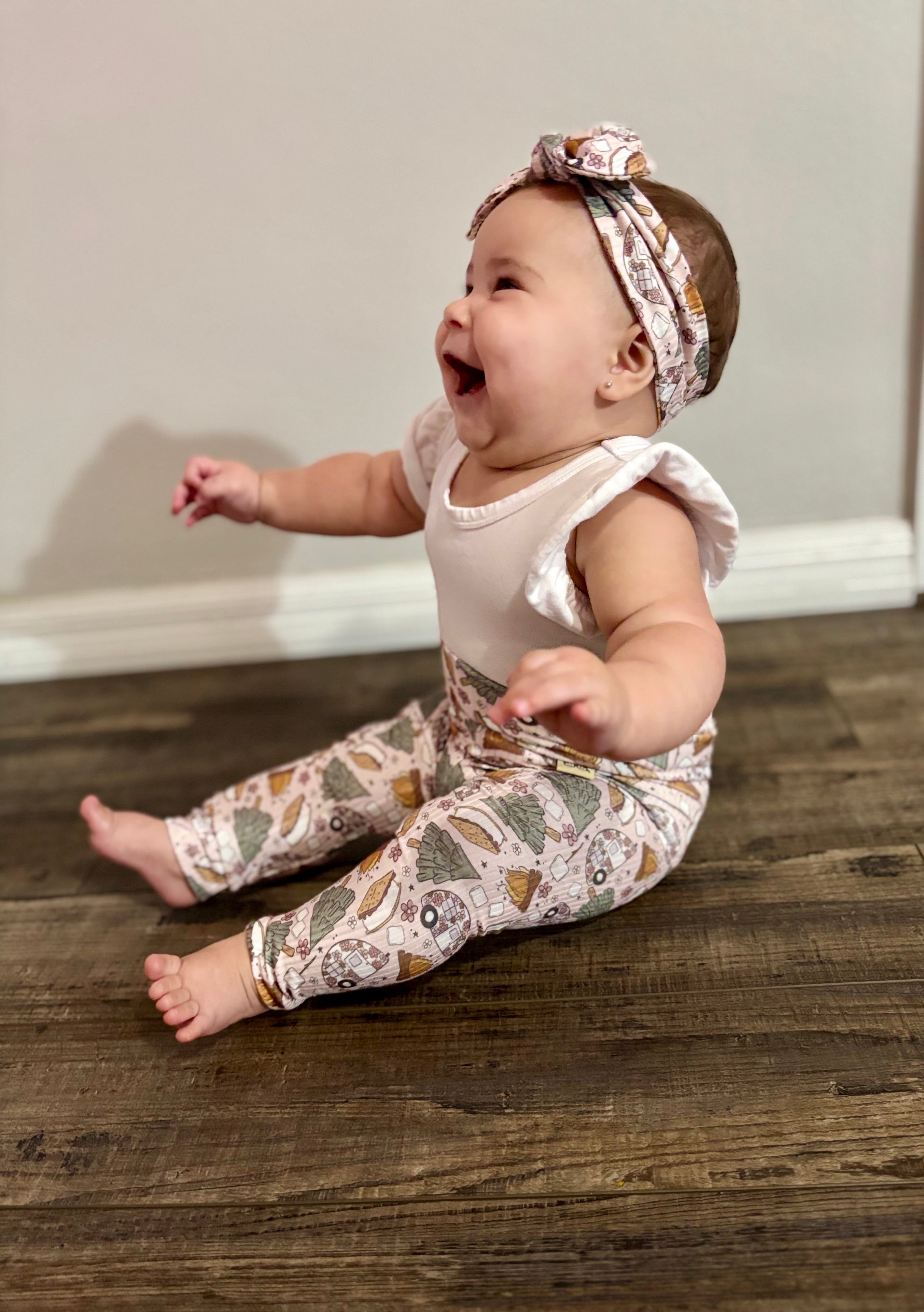 Baby sitting on a wooden floor wearing a white top and matching camping print leggings and headband.