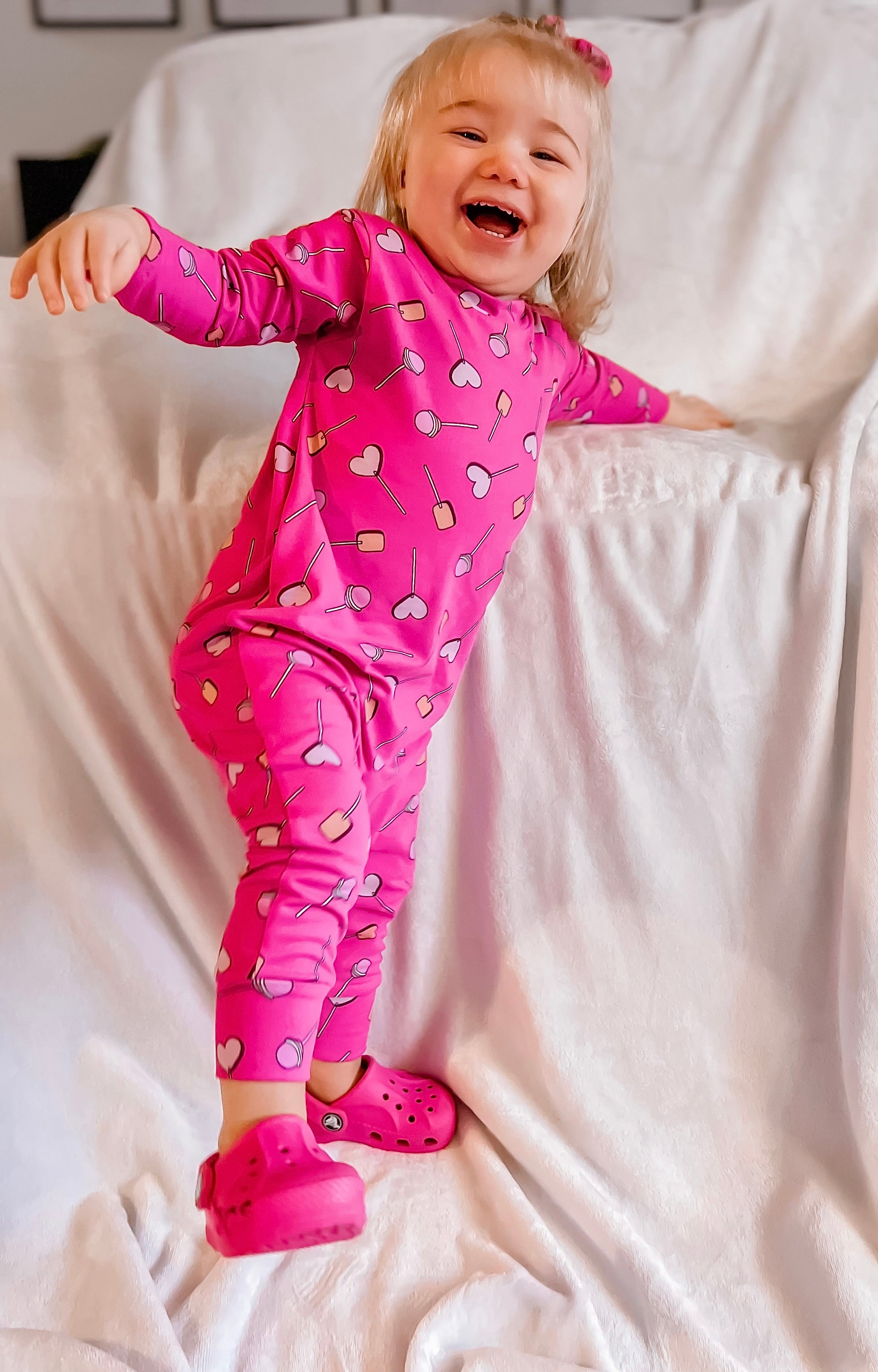 Smiling toddler wearing a handmade bright pink outfit with heart and lollipop patterns, featuring a soft and stretchy two-piece design. Perfect for toddlers, this cozy and playful set is ideal for everyday wear or special occasions. Shop unique baby and toddler clothing today!