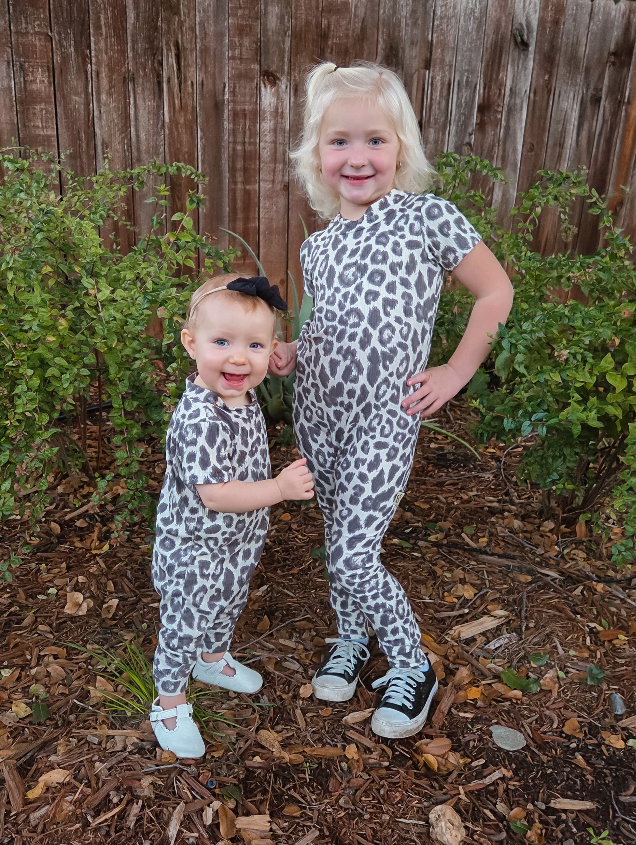 Two children in matching leopard print outfits standing outdoors with a wooden fence and greenery in the background.