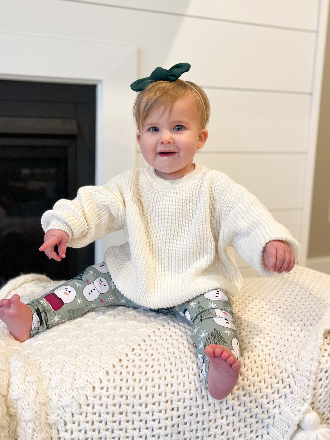 Baby sitting on a white surface wearing a white sweater and patterned pants with a green bow in hair.