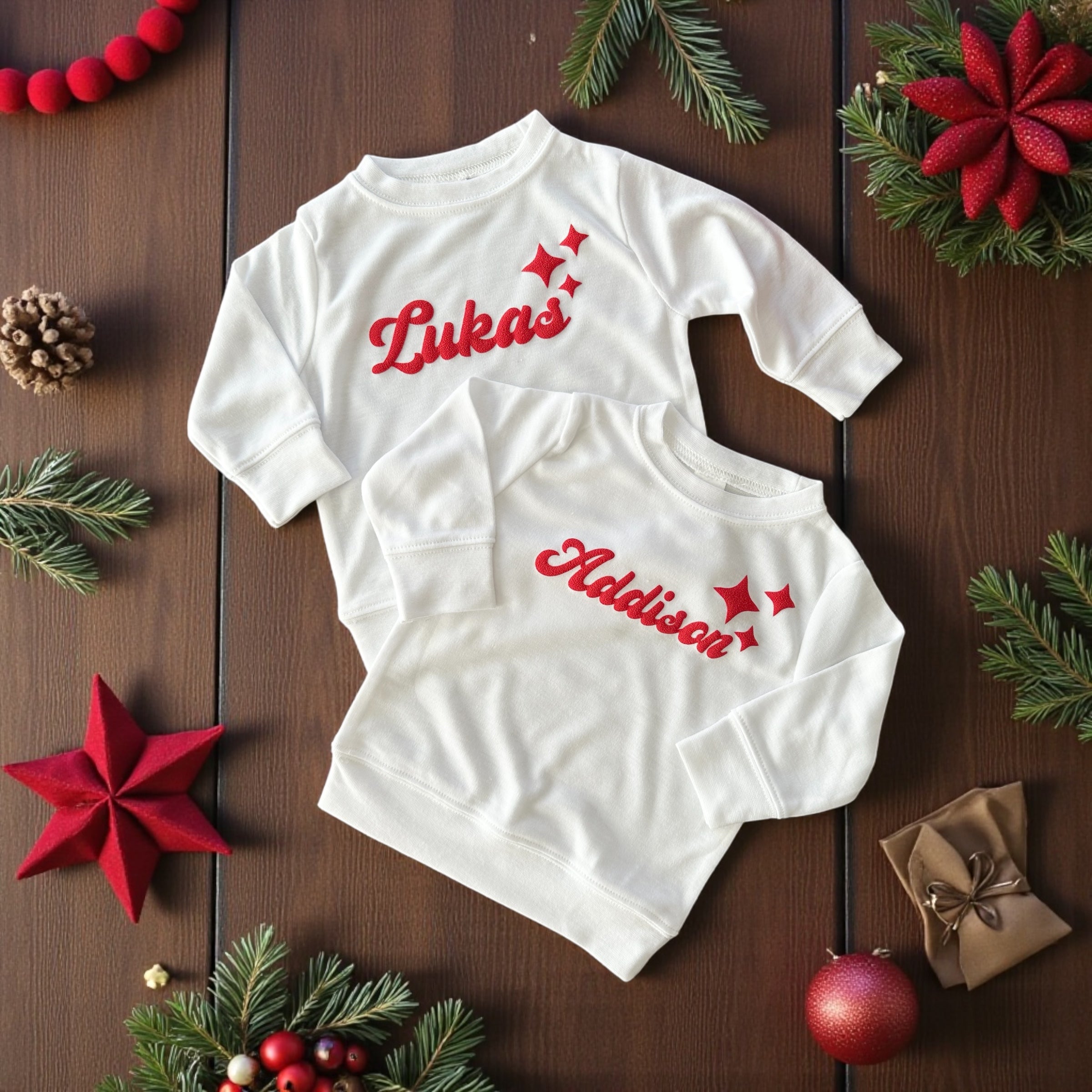 Two white baby sweatshirts with red personalized names “Lukas” and “Addison” and star accents, styled with Christmas decorations.