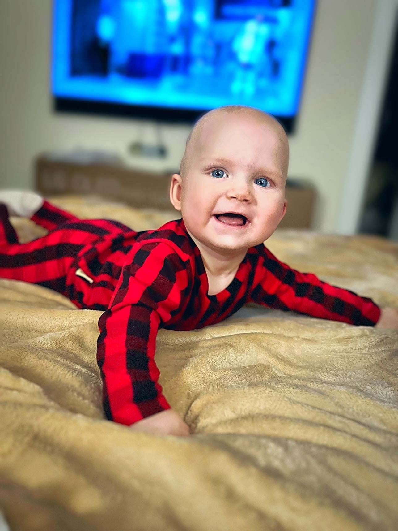 Smiling baby in a red buffalo plaid outfit, lying on a cozy beige blanket, radiating holiday cheer.
