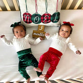 Twin babies lying on a festive-themed crib setup, one wearing emerald green velvet leggings and a matching headband, and the other in red velvet leggings with a red bow. Celebrating their 3-month milestone in holiday style.