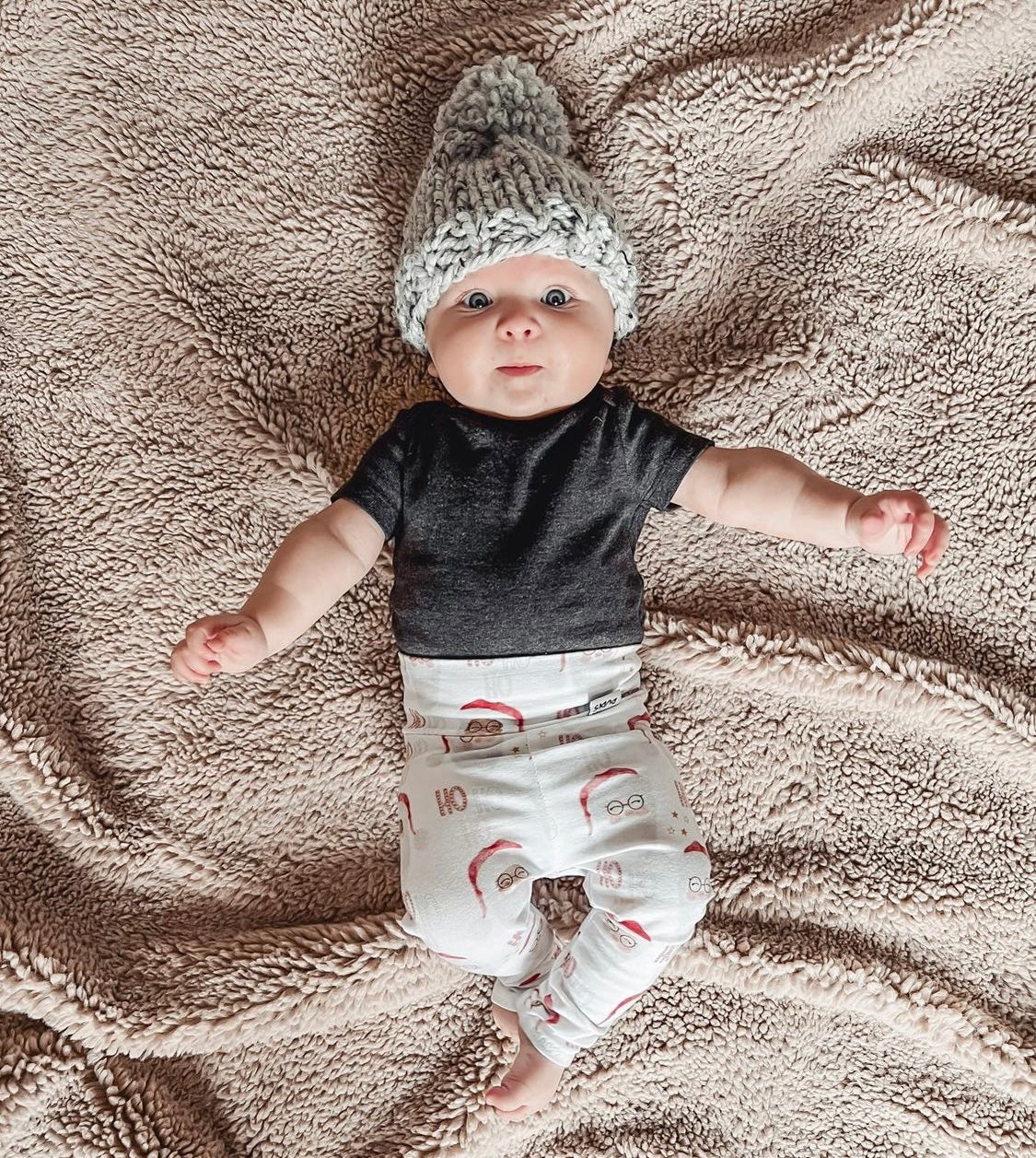 Baby wearing Vintage Santa leggings with festive red and white print, styled with a black top and a cozy gray knit hat, lying on a soft beige blanket. Perfect holiday outfit for babies and toddlers.