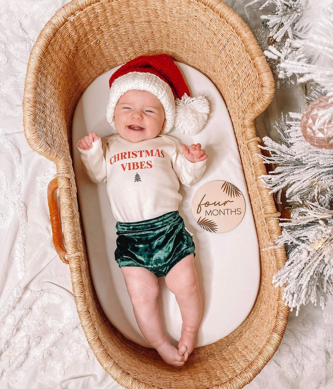 Smiling baby dressed in emerald green velvet bummies and a Santa hat, lying in a wicker bassinet with "Christmas Vibes" onesie. Perfect for festive moments.