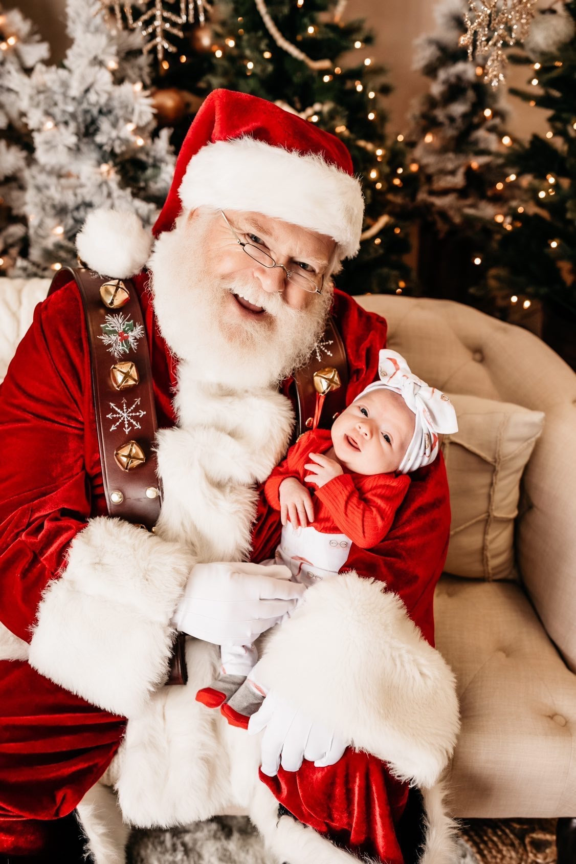 Smiling Santa Claus holding a baby dressed in a festive red outfit, Vintage Santa leggings, and a matching top knot headband. Set against a backdrop of beautifully decorated Christmas trees and warm holiday lights. Perfect holiday moment captured with seasonal charm.