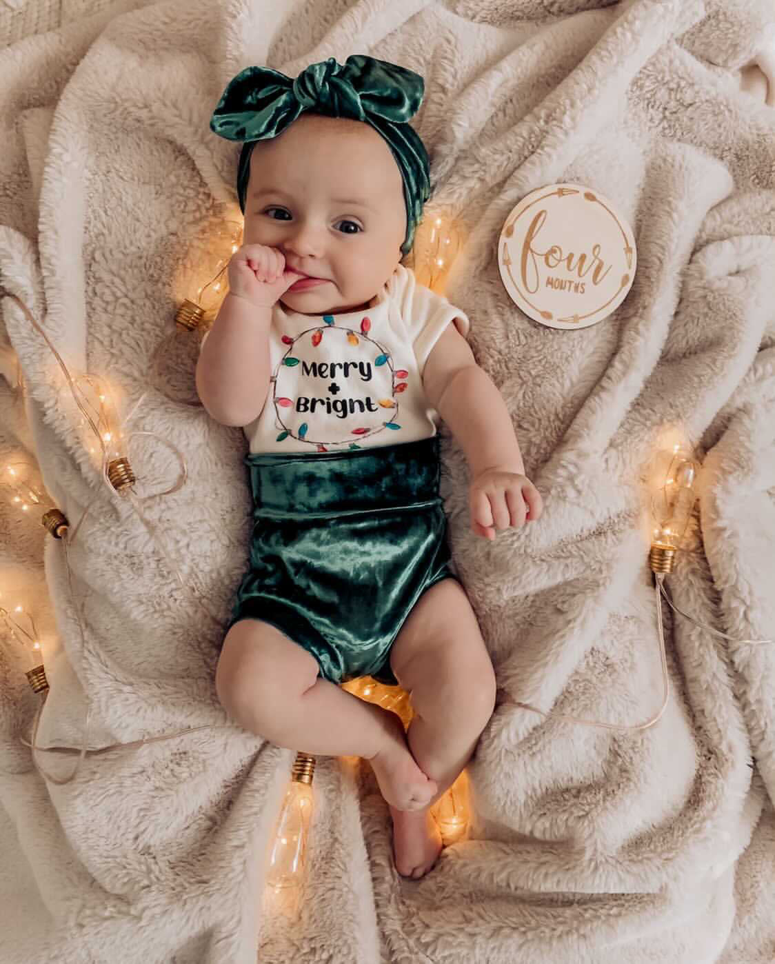 Baby lying on a cozy beige blanket surrounded by Christmas lights, wearing emerald green velvet bummies, a matching headband, and a "Merry & Bright" bodysuit. A sweet holiday look.