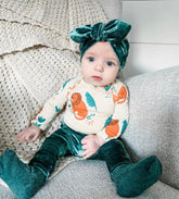 Baby sitting on a neutral-toned couch wearing emerald green velvet leggings, a matching headband, and a colorful lion print bodysuit. A perfect combination of luxury and playful style.