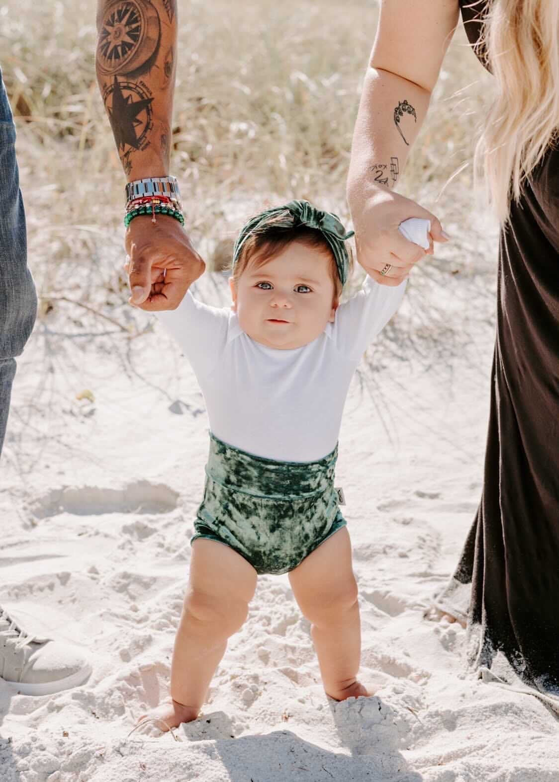 Baby wearing emerald green velvet bummies and matching headband, supported by parents, standing on white sand beach. A chic holiday or special occasion outfit.
