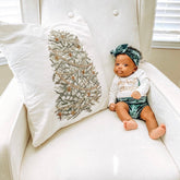 Newborn baby dressed in emerald green velvet bummies with a matching headband, lounging on a white chair next to a festive Christmas tree pillow. Perfect for first holiday photos.