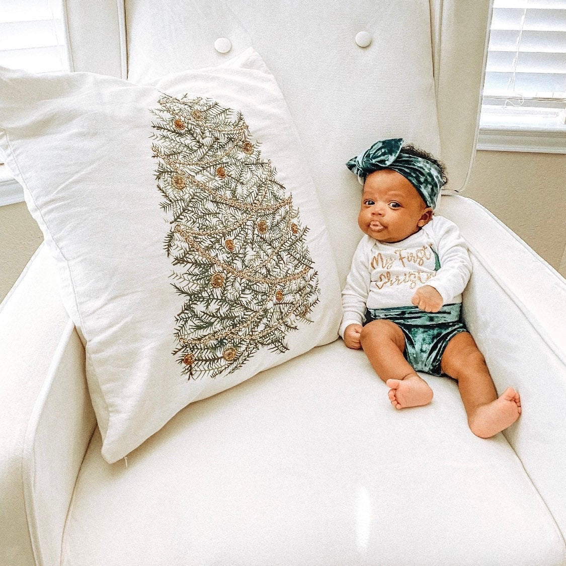 Newborn baby dressed in emerald green velvet bummies with a matching headband, lounging on a white chair next to a festive Christmas tree pillow. Perfect for first holiday photos.
