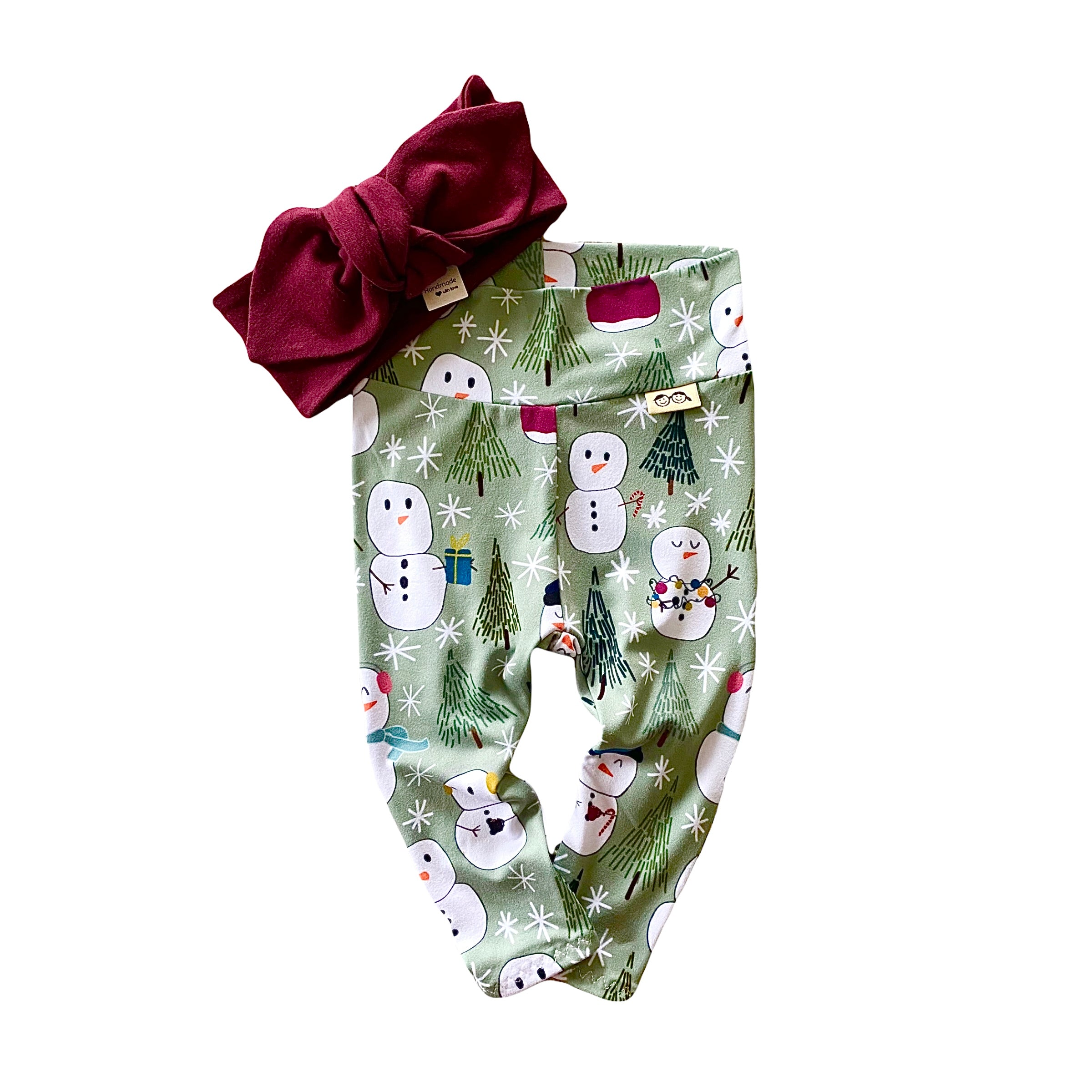 Snowman Print Leggings featuring a playful snowman and winter tree design on a soft green background, paired with a burgundy bow headband. Handmade for babies and toddlers, perfect for holiday celebrations and seasonal outfits.