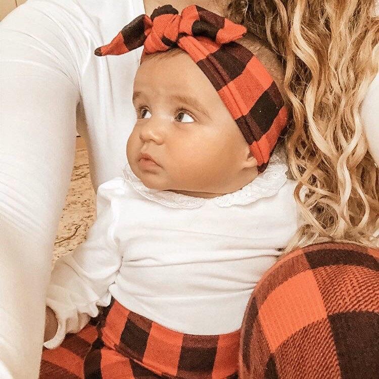 Baby wearing a red buffalo plaid headband and matching pants, sitting with mom in a cozy holiday outfit.
