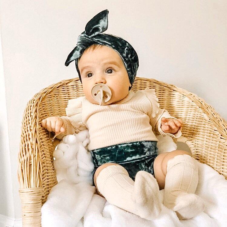 Baby sitting in a wicker chair wearing emerald green velvet bummies, a matching headband, and cream-colored socks and top. A stylish outfit for everyday or special occasions.