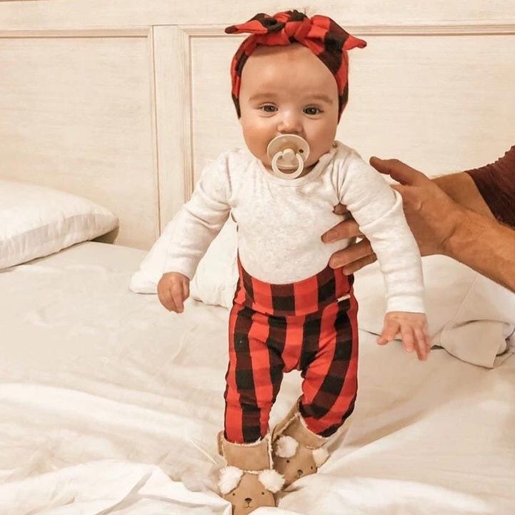 Sweet baby standing with support, dressed in a cozy red buffalo plaid headband and leggings, paired with a white onesie and fuzzy reindeer booties