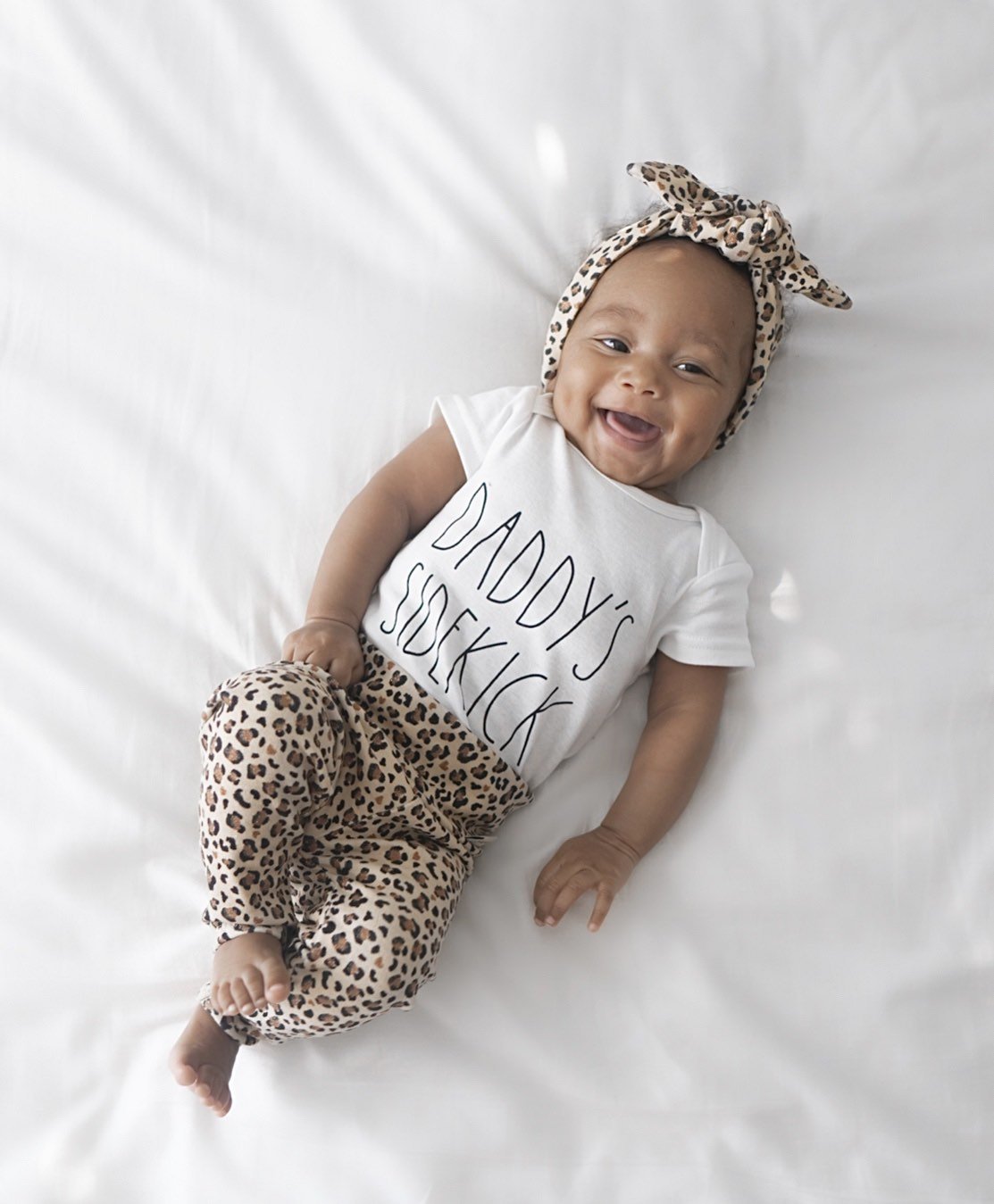 Smiling baby lying on a white blanket wearing leopard print leggings paired with a matching leopard headband and a "Daddy&