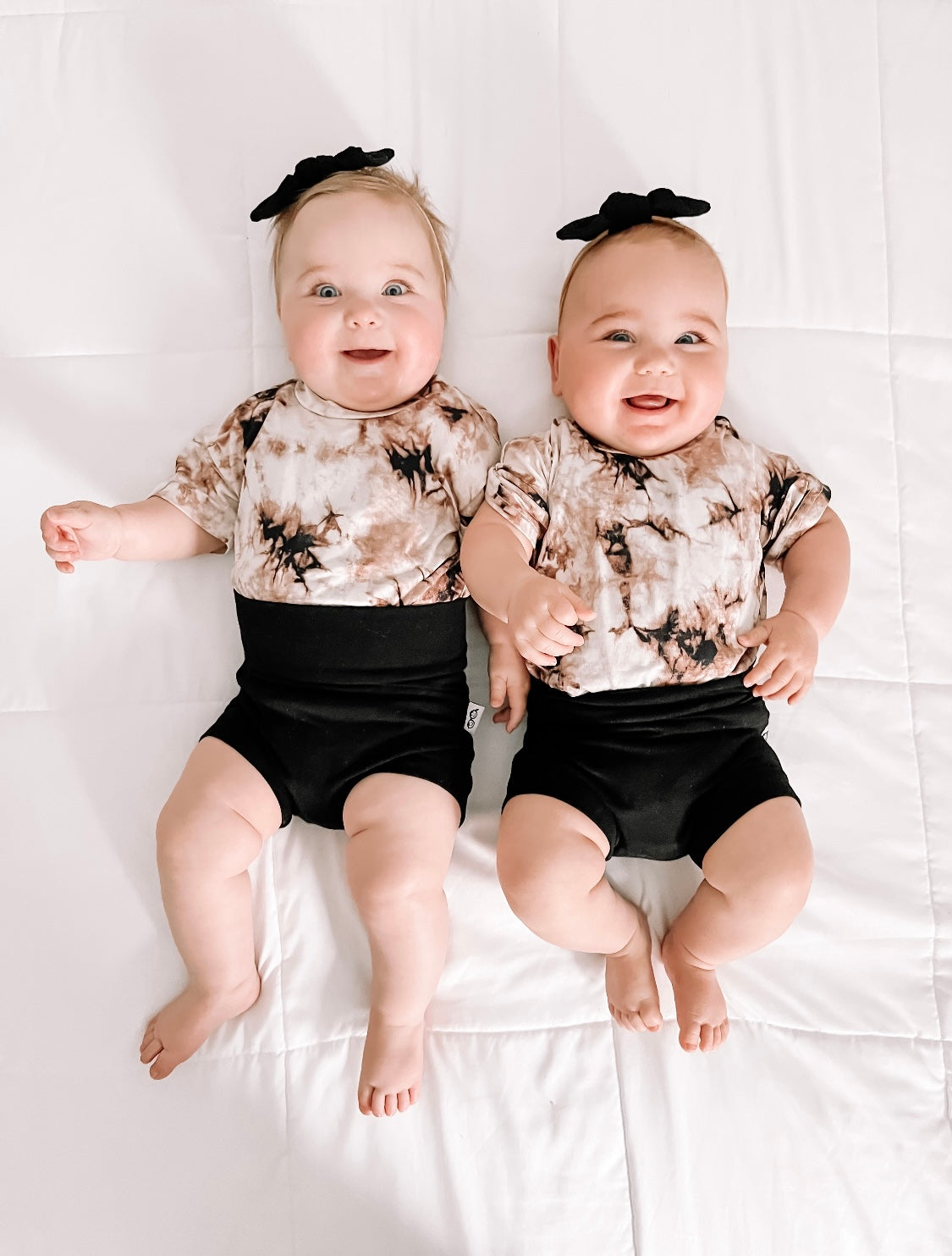 Two smiling babies lying down wearing black bummies and headbands with a tie-dye pattern.