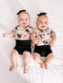 Two smiling babies lying down wearing black bummies and headbands with a tie-dye pattern.