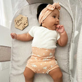 Baby in a crib wearing a white shirt and peach shorts with a headband, next to a &