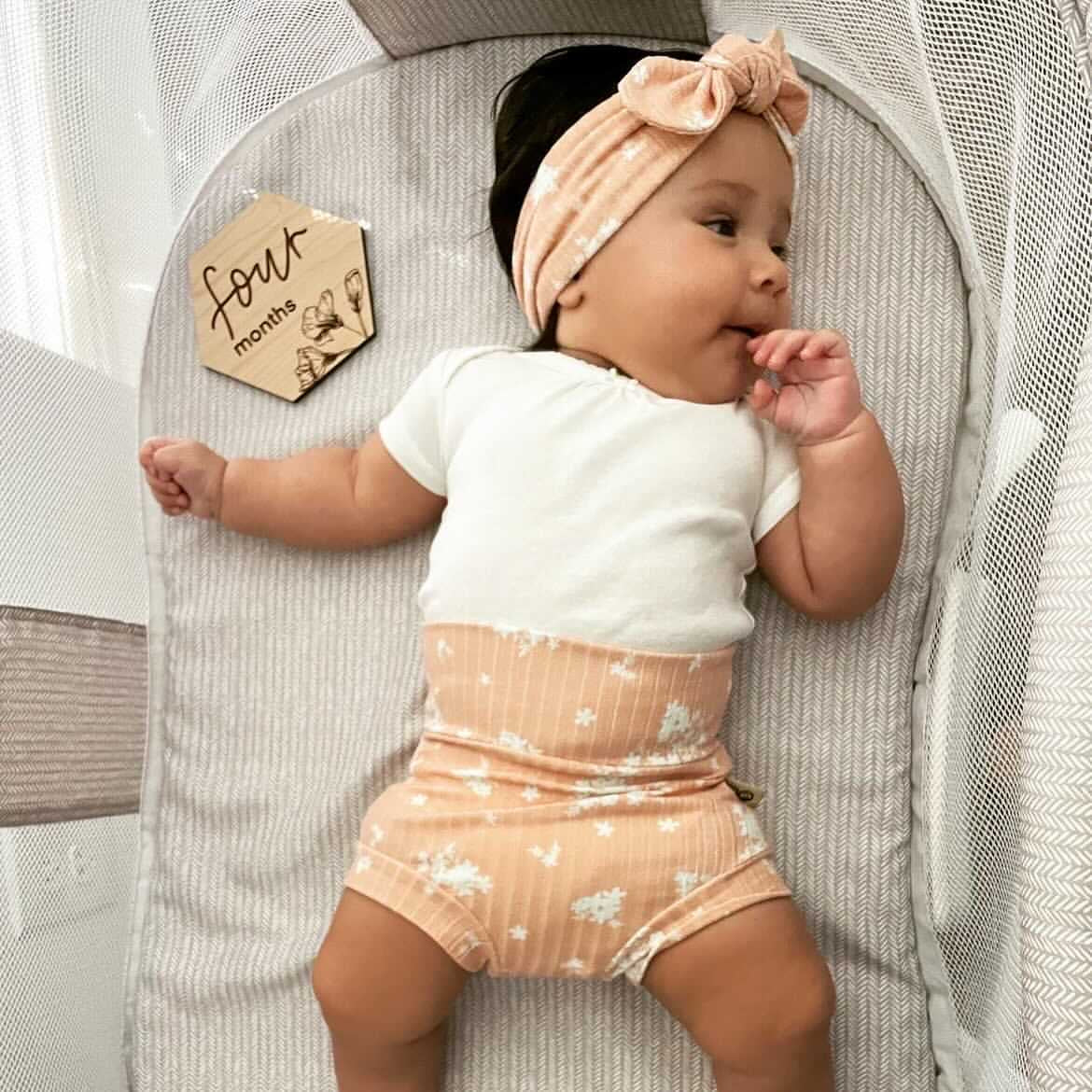 Baby in a crib wearing a white shirt and peach shorts with a headband, next to a &