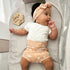Baby in a crib wearing a white shirt and peach shorts with a headband, next to a &