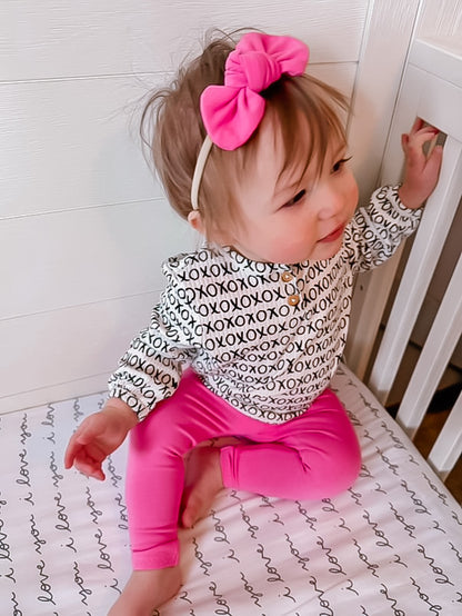 Baby girl wearing hot pink leggings and an XOXO print long-sleeve top, paired with a matching pink bow headband. A trendy and comfortable baby outfit, ideal for casual wear, playtime, or special occasions. Handmade leggings available in sizes preemie to 4T.