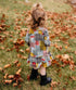 A little girl standing outdoors in a Retro Plaid Dress with a ruffled hem, surrounded by autumn leaves for a charming fall look.