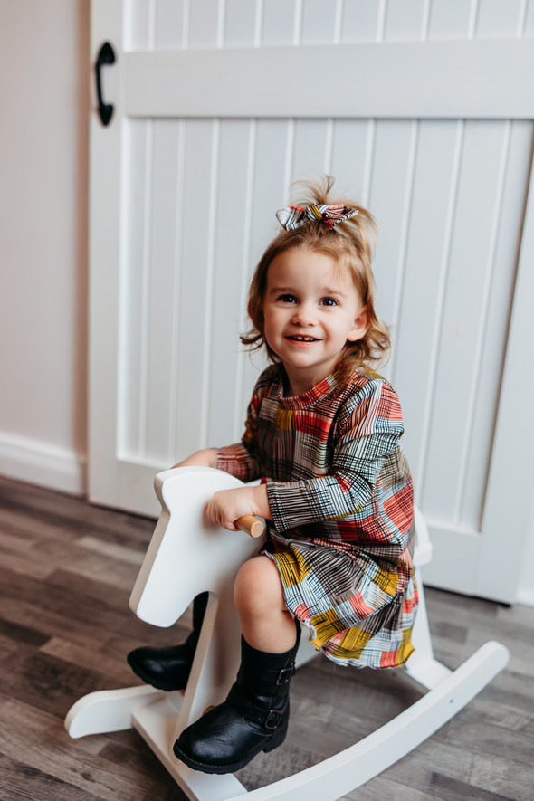 A smiling toddler sitting on a white rocking horse indoors, wearing a Retro Plaid Dress paired with black boots and a matching plaid bow.&quot;