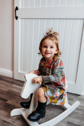 A smiling toddler sitting on a white rocking horse indoors, wearing a Retro Plaid Dress paired with black boots and a matching plaid bow."