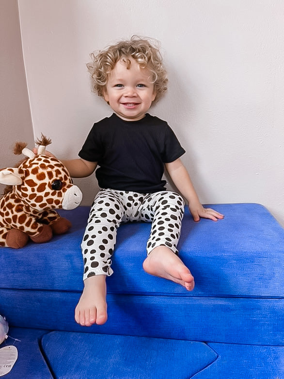 Smiling toddler wearing white and black polka-dot baby leggings paired with a black top, sitting on a blue play couch with a giraffe plush toy. Handmade, comfy, and stylish leggings perfect for infants and toddlers.