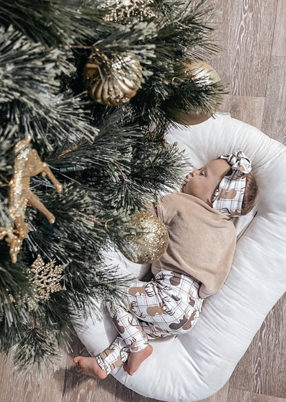 Baby peacefully sleeping under a Christmas tree, dressed in reindeer plaid leggings and a matching headband, perfect for holiday photos.