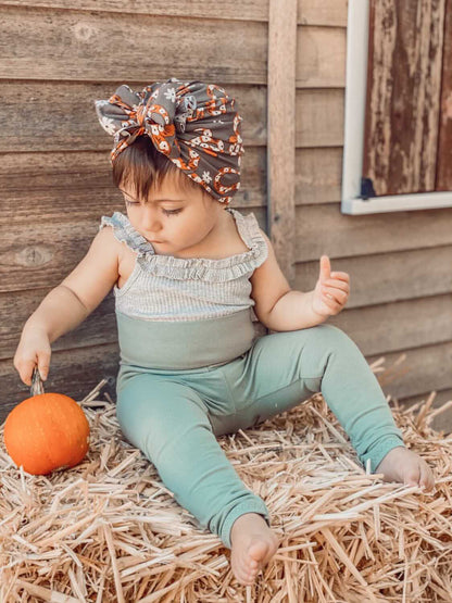 Child sitting on hay with a pumpkin, wearing a headband and green outfit, against a wooden wall.