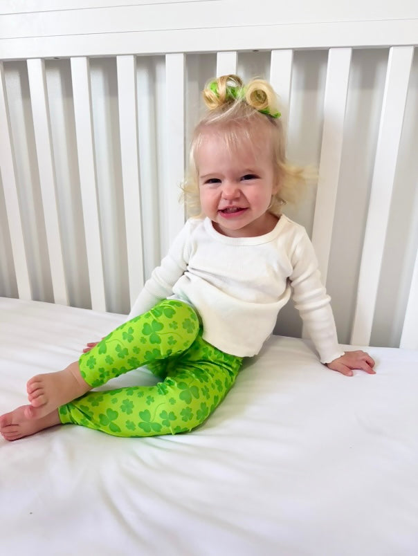 Toddler girl wearing green shamrock leggings for St. Patrick’s Day – Festive clover print leggings paired with a white long-sleeve top. Perfect for lucky little ones celebrating St. Paddy’s Day in style. Soft, stretchy fabric for all-day comfort. 