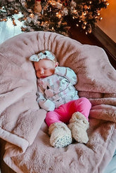 Newborn baby peacefully sleeping under a decorated Christmas tree, wearing a festive white and black pine tree print outfit, paired with bright pink pants, a matching headband, and cozy beige booties. A perfect holiday moment captured with seasonal charm.