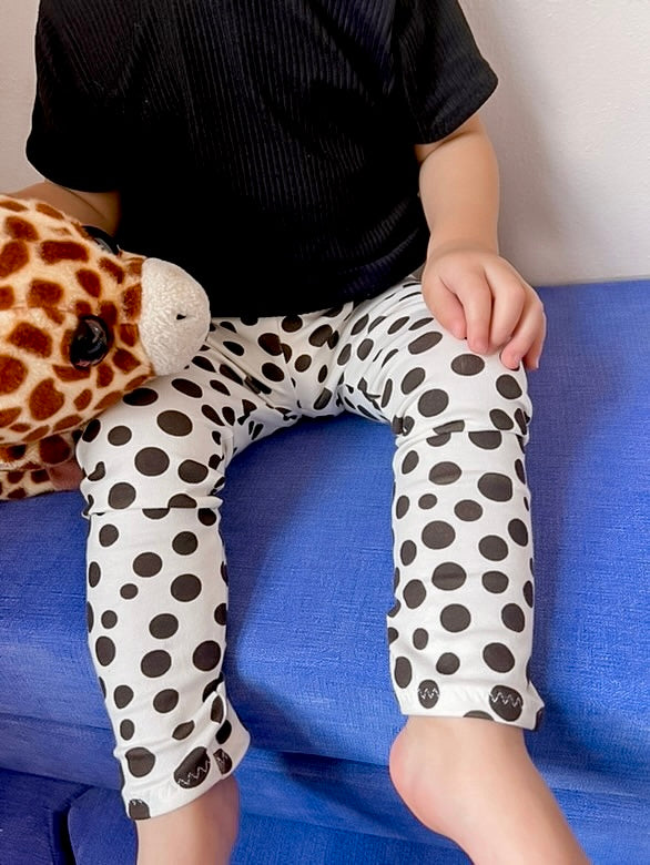 Close-up of white baby leggings with black polka dots, shown on a toddler seated on a blue couch. Soft, stretchy, and perfect for everyday wear or gifting, these handmade baby pants are ideal for boys and girls.