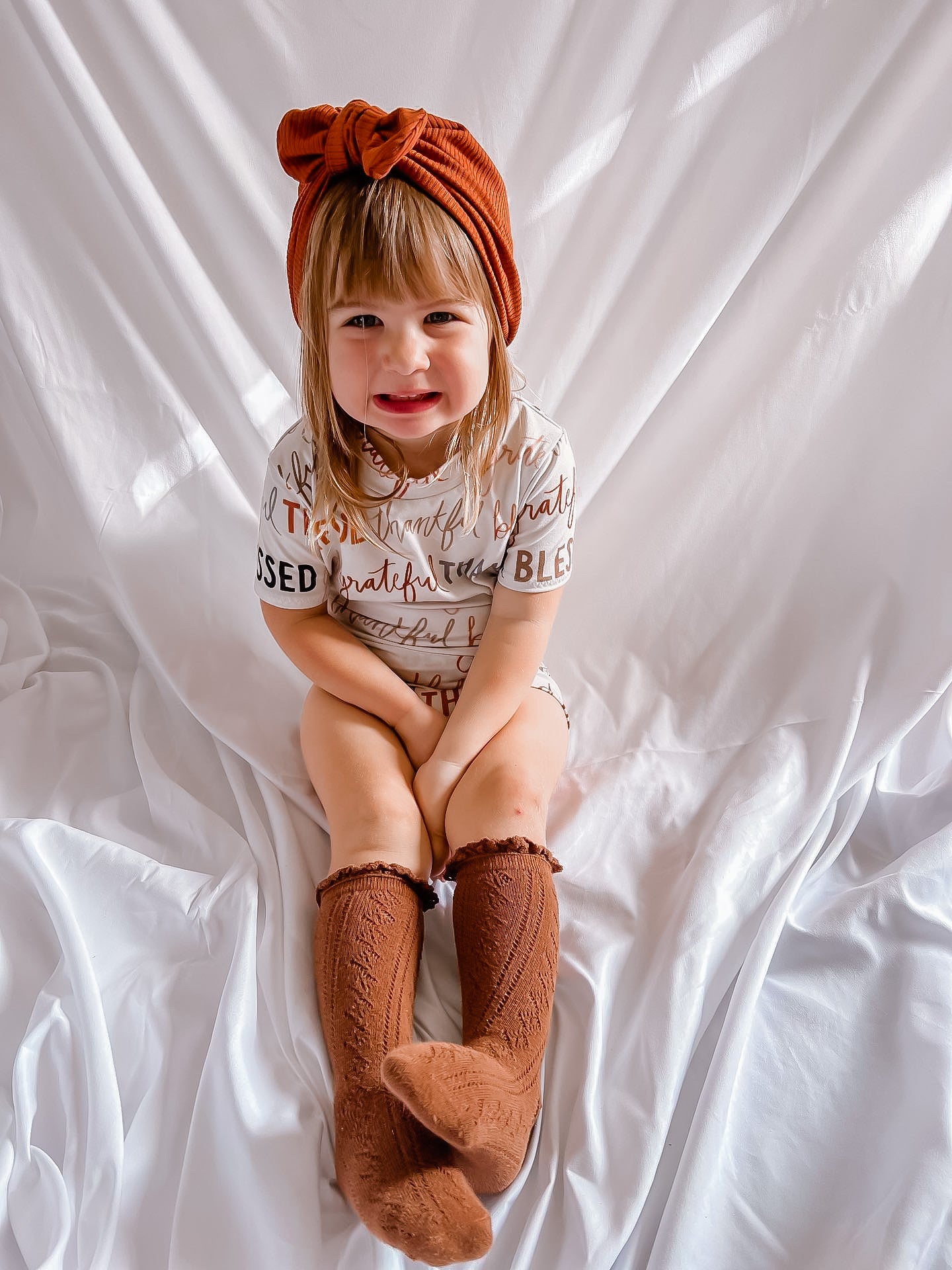 Child wearing a white shirt with text, brown knitted socks, and an orange headband on a white background