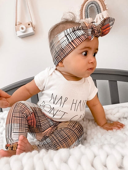 Baby wearing a white onesie with text and plaid pants, sitting on a white surface.