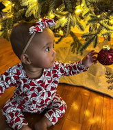 Baby dressed in a cozy Santa Hats romper sitting under a Christmas tree, reaching for a red ornament with warm holiday lights glowing in the background.