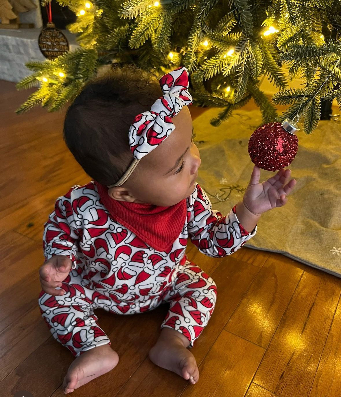 Baby dressed in a cozy Santa Hats romper sitting under a Christmas tree, reaching for a red ornament with warm holiday lights glowing in the background.