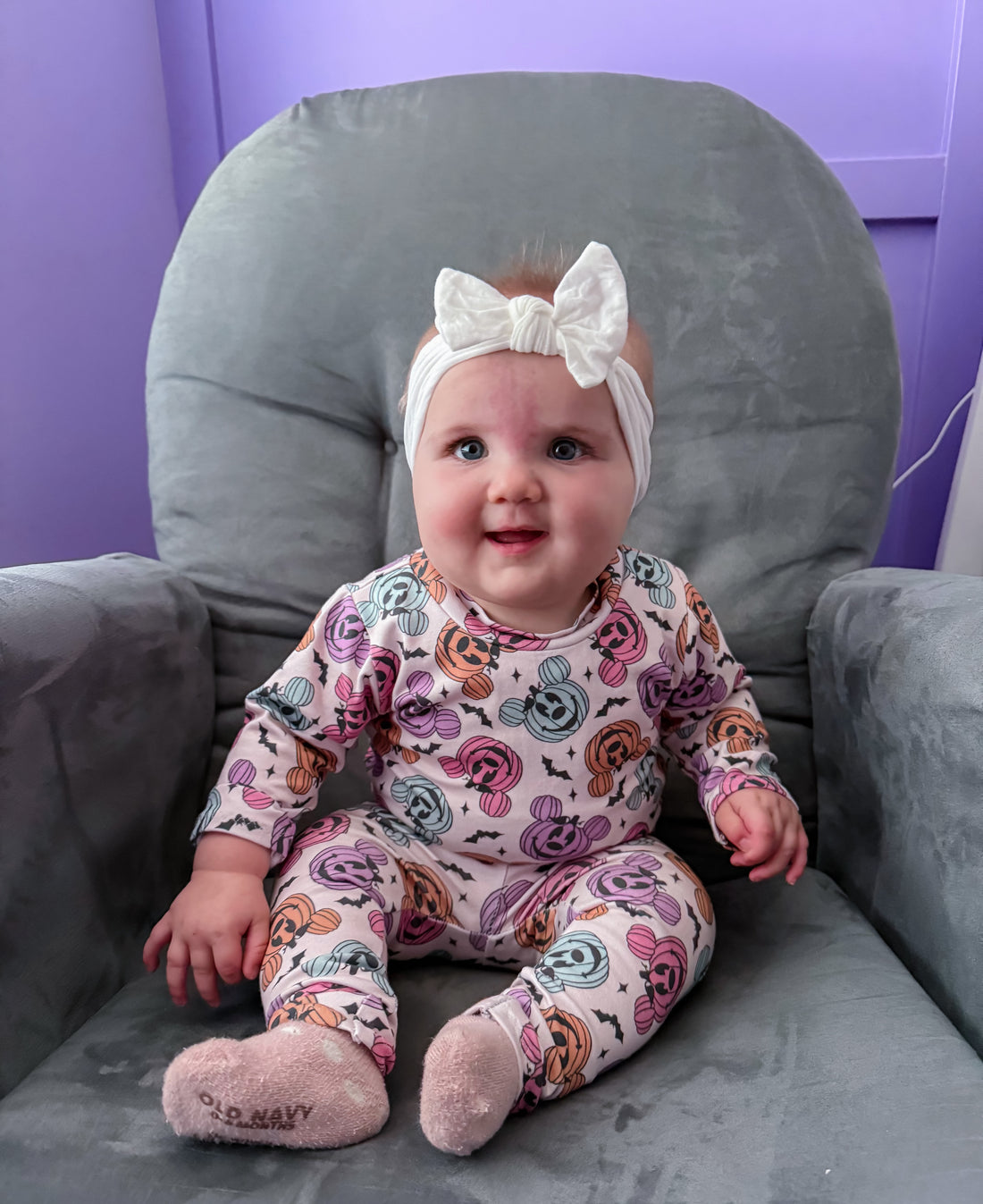 Baby sitting on a gray chair wearing a colorful onesie and white headband.