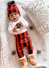 Baby dressed in cozy red buffalo plaid leggings and matching beanie, lying on a chunky knit blanket holding a pinecone, surrounded by winter decor.