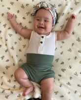 Baby wearing a white and green outfit with a floral headband on a patterned blanket