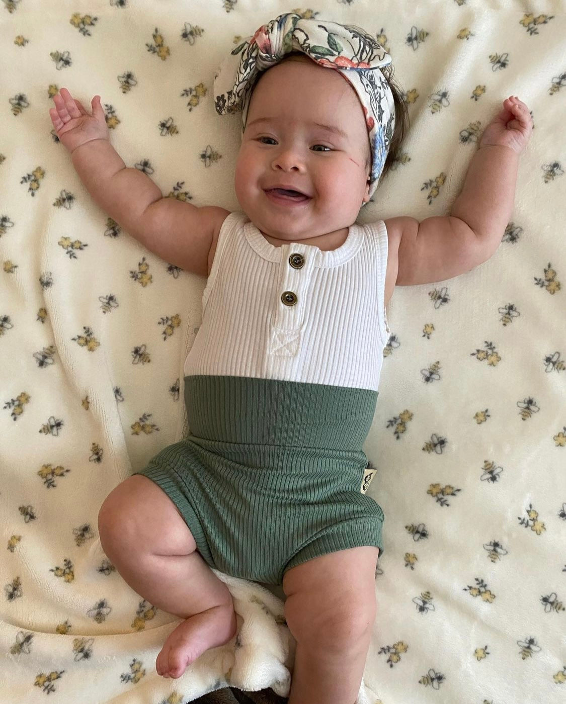 Baby wearing a white and green outfit with a floral headband on a patterned blanket