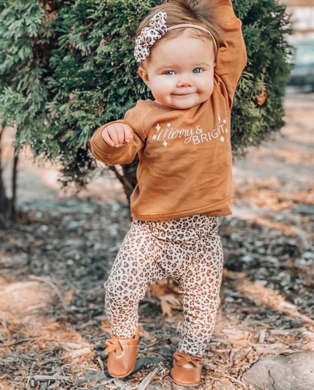 Adorable toddler standing outdoors in leopard print leggings, a brown &quot;Merry &amp; Bright&quot; sweater, and tan bow moccasins, accessorized with a matching leopard print bow headband.