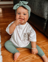 Smiling baby wearing ribbed green leggings, a matching headband, and a floral-pocket white tee, sitting on a wooden floor.