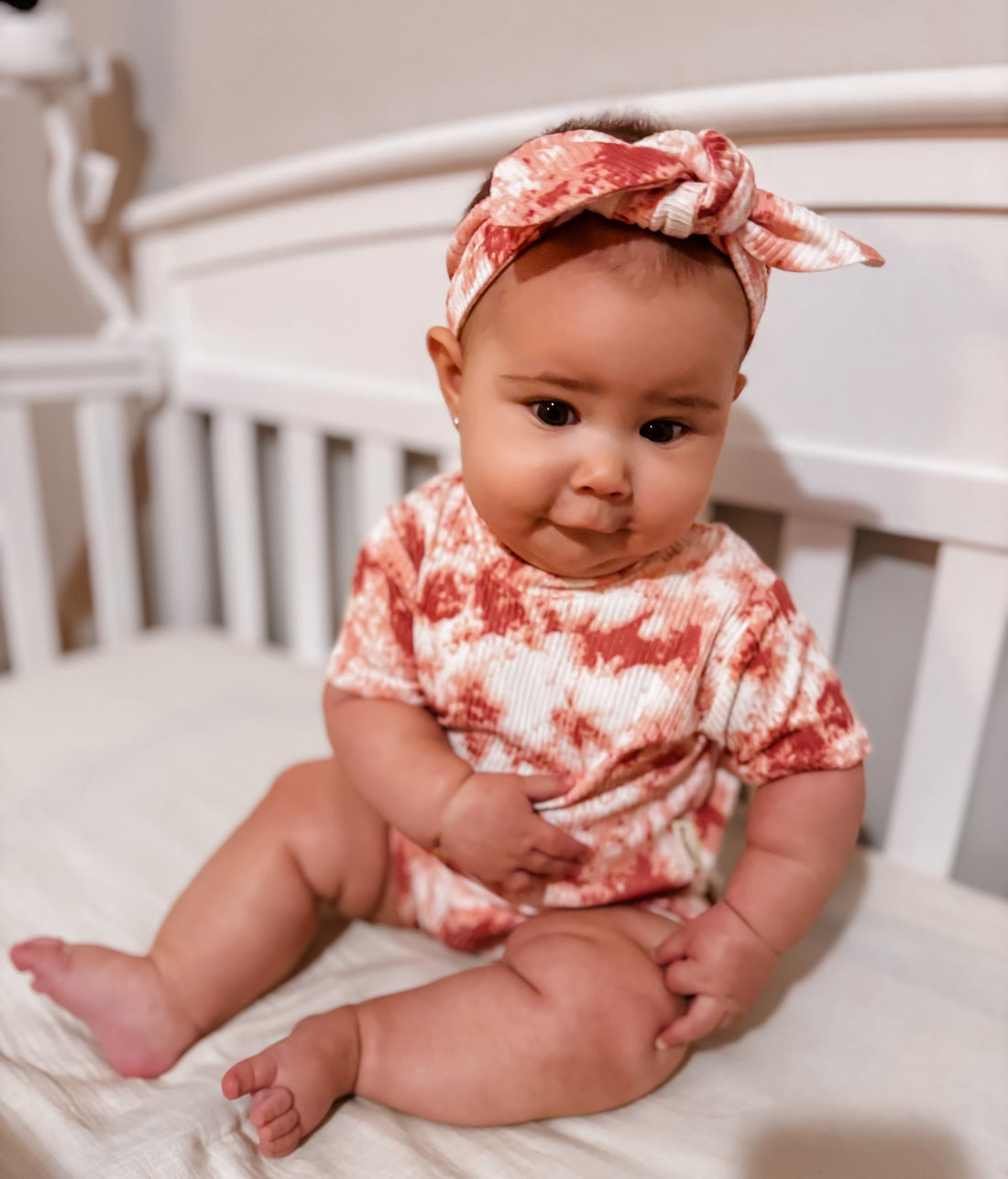 Baby wearing a red and white patterned outfit with a matching headband, sitting in a crib.